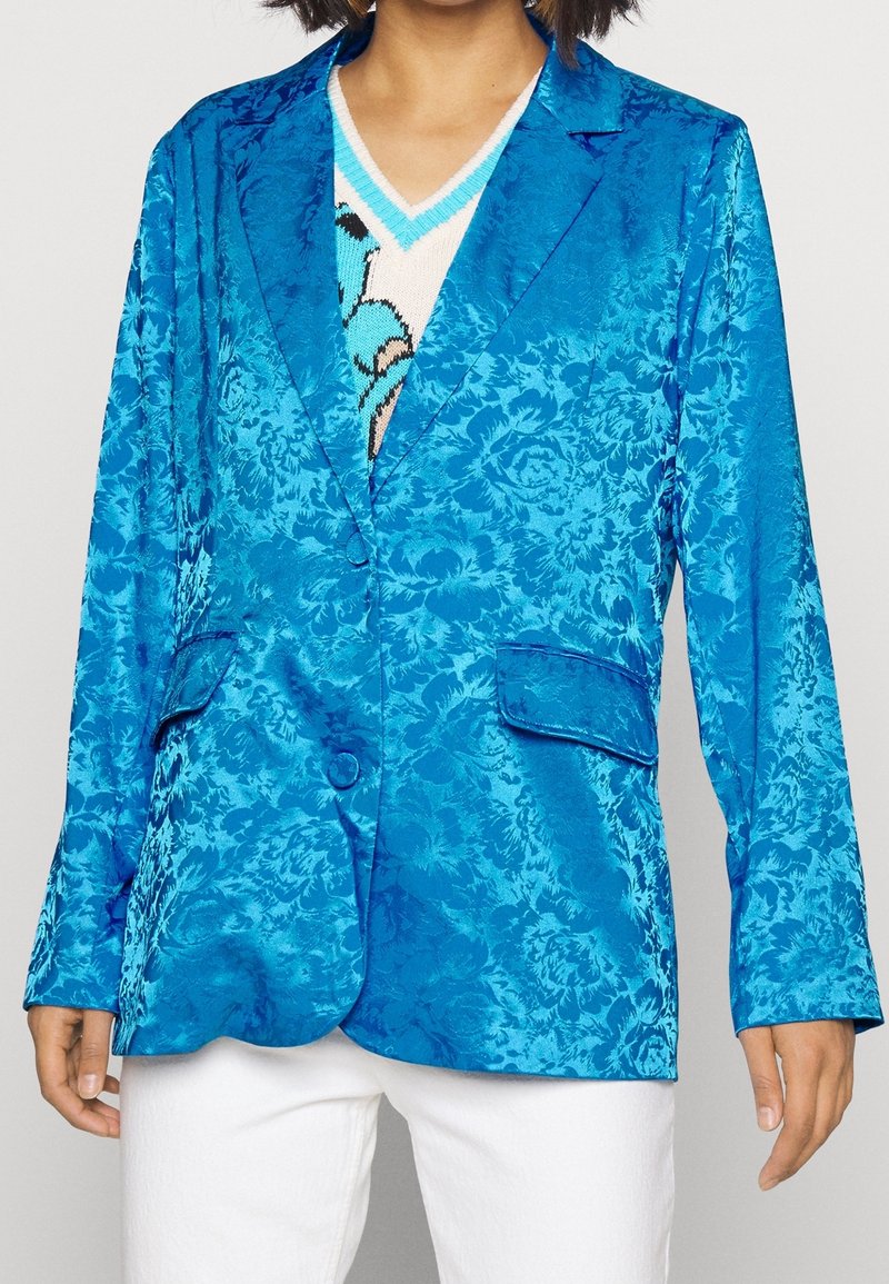 Femme portant un blazer jacquard floréal bleu vif sur un pullover à col en V à motifs et un pantalon blanc, visible des épaules jusqu'aux hanches.