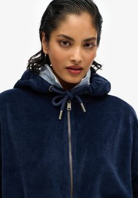 Sudadera de terciopelo azul marino con cierre de cremallera, capucha con cordón y detalles metálicos, y un sutil logo bordado en el pecho.