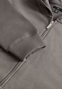 Grauer Kapuzenpullover mit Reißverschluss, geripptem Bündchen und Metall-Reißverschlussschieber, der Nahaufnahmen vom Ärmel und oberen Vorderteil zeigt.