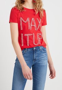 Camiseta de algodón roja con letras en relieve en blanco que dicen "MAX ITUR". Combinada con jeans de mezclilla azules, la camiseta tiene un escote redondo y mangas cortas.