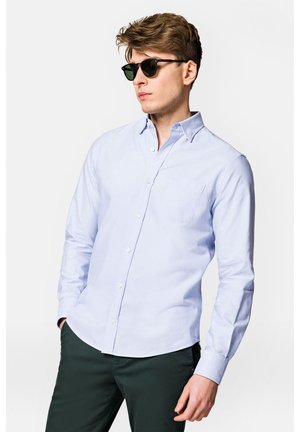 Jasnoniebieska koszula z długim rękawem, z pojedynczą kieszenią na piersi i miękką bawełnianą fakturą. Noszona z ciemnozielonymi spodniami i czarnymi okularami przeciwsłonecznymi.