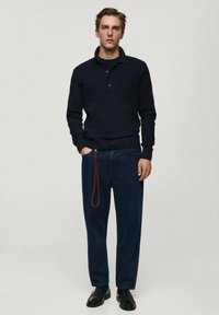 Navy ribbed long-sleeve polo shirt with a collar, paired with loose-fitting dark denim jeans featuring a red side loop. Black shoes.