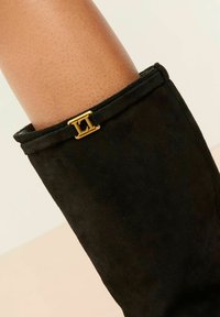 Stivaletto in suède nero con un emblema dorato vicino alla parte superiore, con un primo piano della pelle con texture sopra. Lo sfondo è neutro e minimalista.