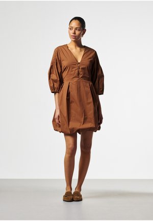 Frau steht und trägt ein braunes, blasenförmiges Kleid mit Puffärmeln und Knopfdetails sowie braune Loafers vor einem einfarbigen Hintergrund.