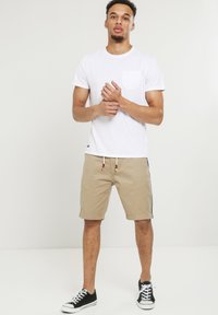 T-shirt blanc à manches courtes avec une poche poitrine associé à un short beige avec un cordon de serrage et des bandes latérales. Porté avec des baskets noires.
