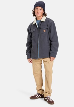 Veste en polaire bleu marine avec un col crème, fermeture éclair, deux poches poitrine, portée avec un pantalon beige et des chaussures marron.