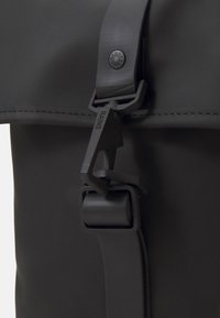 Primo piano di una borsa impermeabile nera, con finitura opaca, tracolla regolabile, chiusura funzionale e dettaglio logo in rilievo.