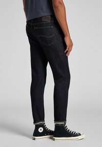 Jeans de mezclilla de lavado oscuro con corte ajustado, que presentan costuras amarillas, cinco bolsillos y una etiqueta de cuero en la cinturilla. Combinados con zapatillas negras de caña alta.