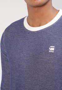 Tmavě modrý svetr s texturou, bílým žebrovaným límcem a malým bílým logem "RAW" na hrudi. Má kulatý výstřih a přiléhavý střih.