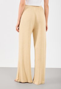 Wijd uitlopende broek in een lichtbeige ribbelstof, met een elastieke tailleband en een zachte textuur. De lengte reikt tot de enkels met een vloeiende val.