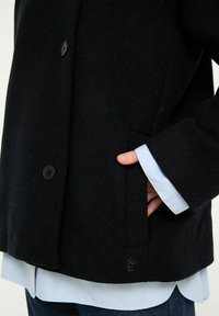 Svart ullblandad jacka med skräddarsydd design, med en enkel ficka, mörka knappar och en ljusblå skjorta under.