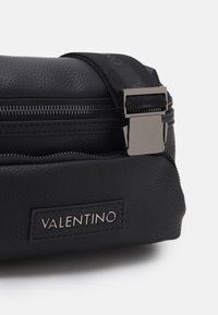 Svart texturerad läderväska med en silvrig dragkedja och en metallspänne. Har en märkesetikett med namnet "VALENTINO."