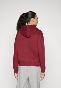 Felpa rossa con cappuccio e cordino, dotata di un ampio cappuccio, con polsini e orlo a costine. Indossata con pantaloni della tuta grigi chiari. Sfondo semplice.