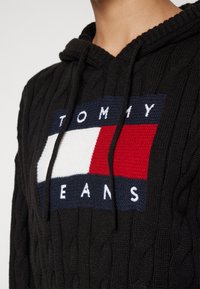 Sort kabelstrikket hættetrøje med snore og et strikket Tommy Jeans-logo i marineblå, hvid og rød på brystet.
