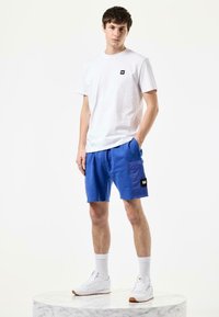 Hvit bomull t-skjorte med lite logo og blå shorts med lomme og merke, kombinert med hvite sneakers og sokker. Stående posisjon.