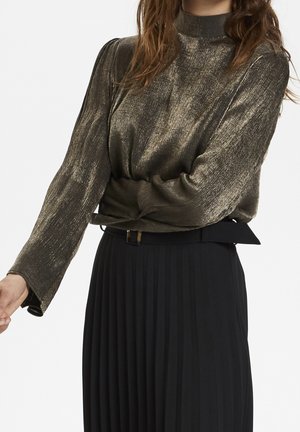 Woman wearing a shiny metallic long-sleeve top tucked into a high-waisted black pleated skirt with a belt.