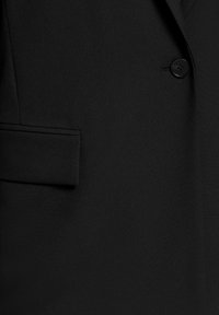 Gros plan sur le tissu noir d'un blazer montrant une poche à rabat à gauche et un seul bouton noir à droite.