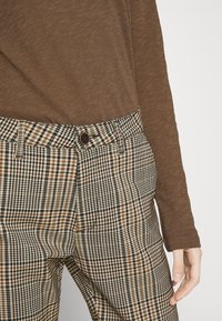 Personne portant un haut à manches longues marron et un pantalon à carreaux beige, noir et marron avec bouton et passants de ceinture, main détendue le long du corps.