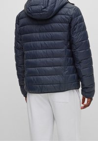 Muž v námořnickém modrém prošívaném kabátu s kapucí a bílých kalhotách, zobrazen zezadu na jednobarevném světlém pozadí.