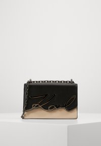 Bolso cruzado de cuero negro y beige con una correa de cadena, que presenta un destacado logo negro en la parte frontal y una forma rectangular elegante.