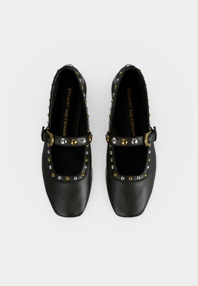 Paire de chaussures Mary Jane noires à bout carré avec des clous dorés et argentés le long des bords et des brides, bouclées sur fond blanc.