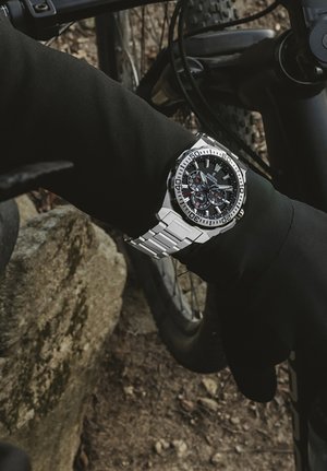 Silberner Edelstahlzeitmesser mit schwarzem Zifferblatt und Chronographenfunktionen, der auf einem schwarzen Handschuh getragen wird, während im Hintergrund ein Mountainbike zu sehen ist.