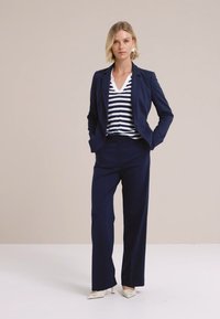 Navy blazer päällystikkuna, jossa on raidallinen tummansininen ja valkoinen yläosa, yhdistettynä tummansinisiin leveälahkeisiin housuihin ja vaaleisiin korkokenkiin; sileä kangas.