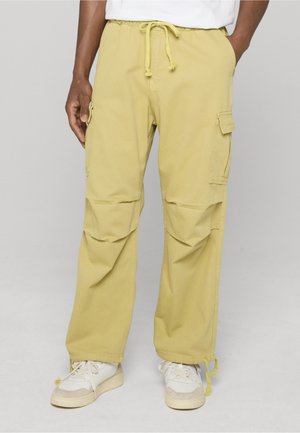 Man wearing yellow cargo pants with drawstrings and white sneakers, standing with one hand in pocket against a plain background.