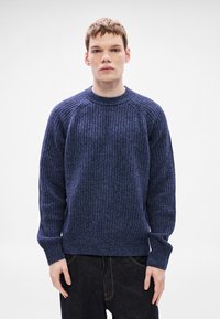 Πλεκτό πουλόβερ σε ναυτικό μπλε με ανάγλυφο μοτίβο, στρογγυλή λαιμόκοψη και ραγκλάν μανίκια. Το ύφασμα φαίνεται μαλακό και ζεστό. Φορεμένο με σκούρο τζιν.