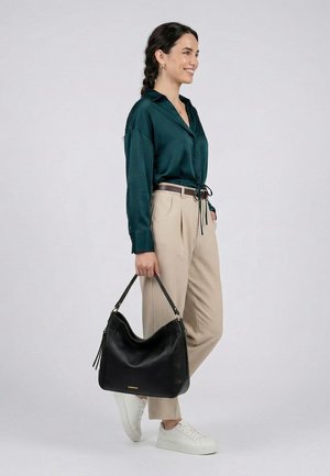 Vrouw staat, draagt een donkergroene blouse, beige broek, witte sneakers, houdt in haar rechterhand een grote zwarte handtas aan de hengsels vast.