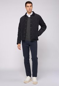 Chaqueta negra acolchada con botones a presión, llevada sobre un suéter gris y pantalones azul marino. Zapatillas blancas completan el atuendo. Diseño y textura simples.