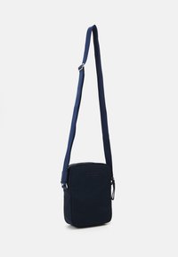 Bolso bandolera en color marino, hecho de tela, de forma rectangular, con correas ajustables, cerrado con una cremallera y con sutiles detalles de costura.