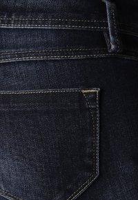 Tela de mezclilla azul oscuro con una textura suave. Presenta un bolsillo trasero, costuras amarillas y un ligero brillo. Diseño clásico de pantalones vaqueros.