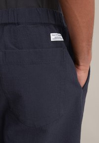 Donkerblauwe broek van textuurstof met een achterzak met een label. De tailleband is elastisch en het materiaal lijkt lichtgewicht.