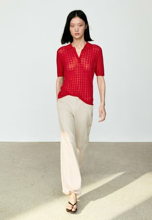 Donna che cammina al chiuso indossando un top a maniche corte rosso in maglia, pantaloni beige a gamba larga e sandali neri su un pavimento chiaro con sfondo semplice.