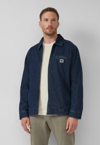 Dunkelblaue Jeansjacke mit einem spitzen Kragen, Reißverschluss, Brusttasche und dezentem Logo-Detail. Tragen über einem hellen Pullover.