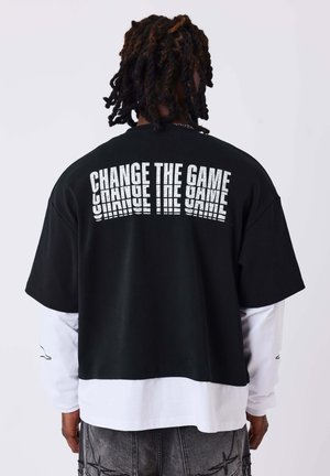 Personne aux cheveux foncés et bouclés porte un t-shirt noir par-dessus des manches longues blanches, avec "CHANGE THE GAME" imprimé en blanc dans le dos.