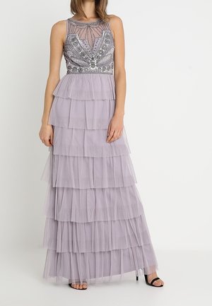 Robe sans manches lavande à volants avec corsage transparent orné, portée par une femme debout, accompagnée de talons ouverts noirs, sur fond blanc.