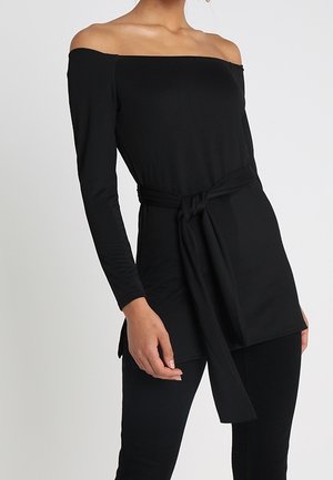 Mujer que lleva una túnica negra de manga larga con los hombros descubiertos y cinturón en la cintura, combinada con pantalones negros ajustados sobre un fondo liso.