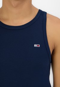 Camică fără mâneci de culoare bleu marin, din bumbac, cu un decolteu rotund și un mic logo brodat cu accente roșii și albe pe piept.
