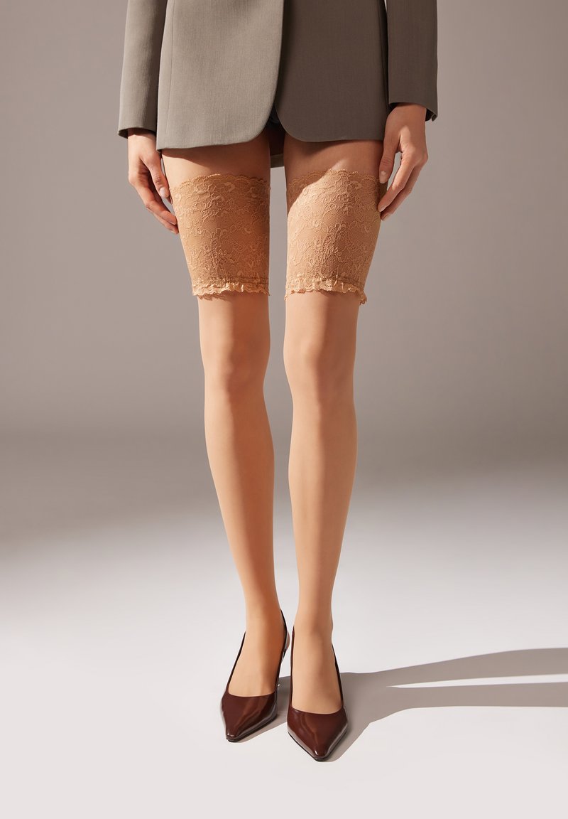 Nude farvede lace thigh-highs med bølgede kanter; parret med spidse brune hæle og stylet under en grå blazer.