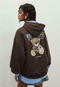 Brauner übergroßer Hoodie mit einem Bärenmotiv auf dem Rücken, versehen mit dem Text "ESPRGO YOURSELF." Darunter trägt er ein hellblaues Hemd.