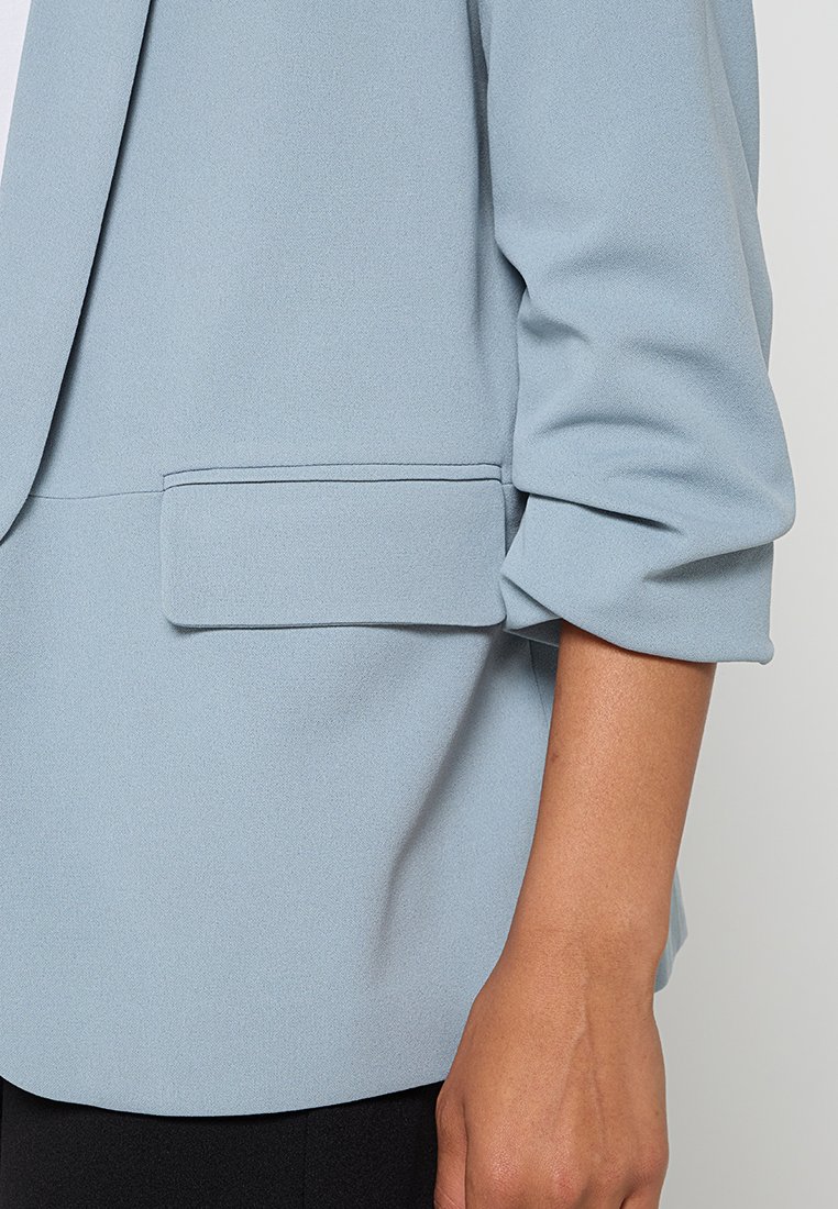 Un blazer bleu clair avec un tissu lisse, un design structuré et une poche poitrine unique. La manche présente un détail froncé.