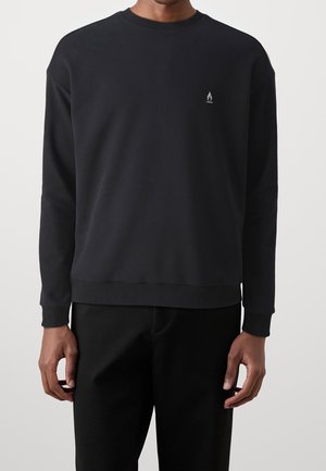 Mann trägt einen schlichten schwarzen Langarm-Sweatshirt mit einem kleinen weißen Flammenlogo auf der linken Brust, kombiniert mit schwarzen Hosen.