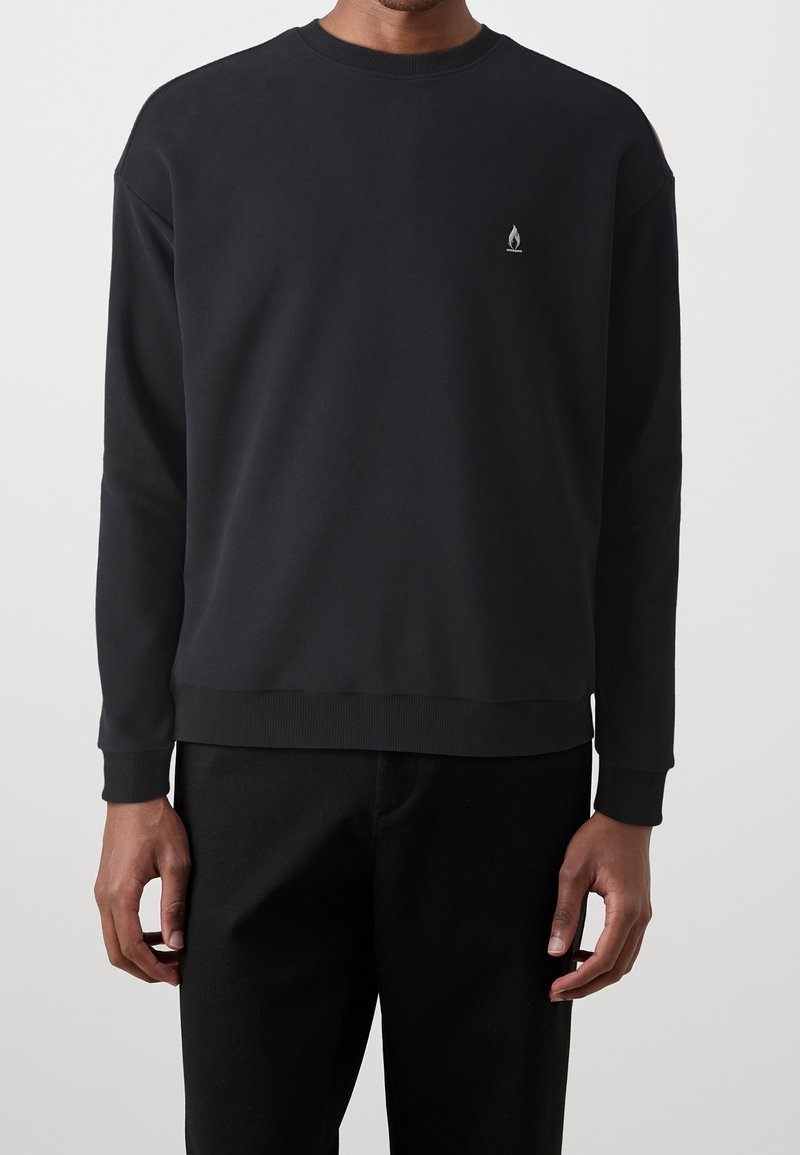 Homme portant un sweat-shirt noir uni à manches longues avec un petit logo de flamme blanche sur la poitrine gauche, associé à un pantalon noir.
