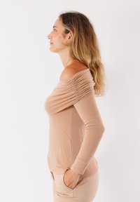 Top a maniche lunghe in tono blush con design off-shoulder e dettagli arricciati, realizzato in morbido tessuto testurizzato. Indossato con pantaloni beige.