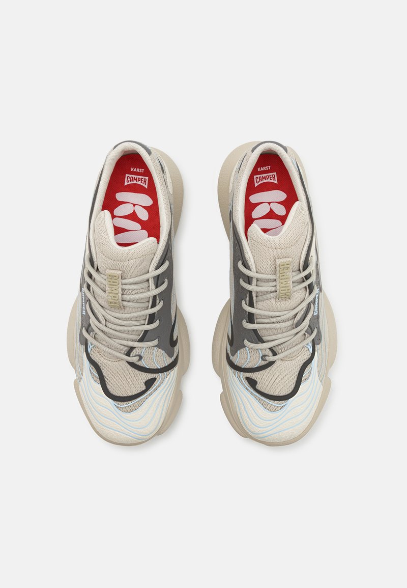 Zapatillas beige y gris con una parte superior de malla texturizada, diseño en capas, cordones prominentes y una plantilla roja con marca blanca.