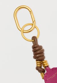 Zelta karabīne un atslēgu gredzens piestiprināts pie loksnes brūnā ādas auklas ar zeltītām gredzenēm un daļēji redzamu purpurkrāsas ādas birku.