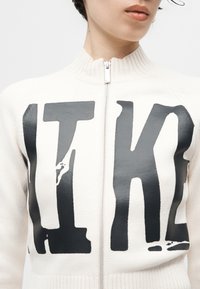 Primo piano di una persona che indossa un maglione bianco con cerniera e grandi lettere nere in grassetto "ITK" sul petto.