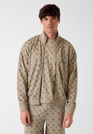 Homme portant une veste beige à fermeture éclair, un T-shirt assorti et un pantalon avec un motif géométrique sombre et des lettres, debout devant un fond uni.